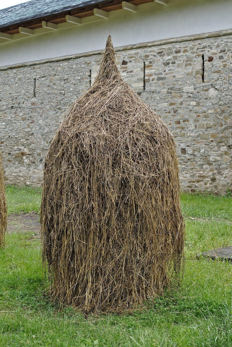 Meule de foin traditionnelle en forme de dôme pointu, faite de brins d’herbe fins et souples, sur une pelouse devant un mur de pierre