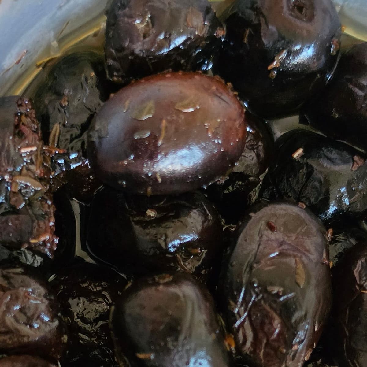 Olives noires en gros plan dans une barquette, brillantes d’huile et parsemées d’herbes, aux teintes allant du noir au brun violacé, certaines lisses, d’autres fripées