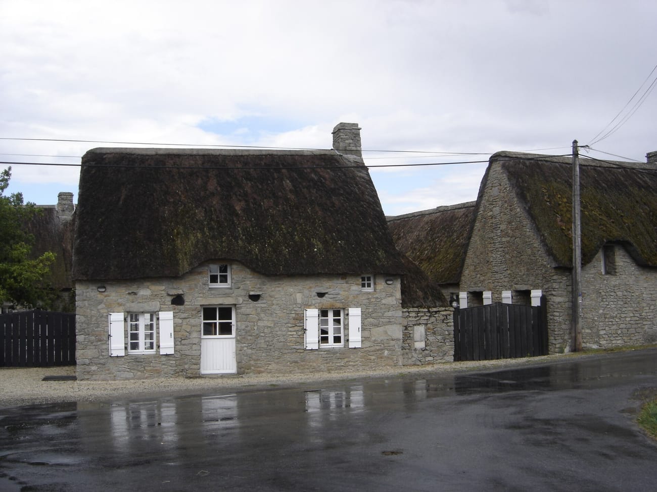 Maison basse en pierre grise coiffée d’un épais toit de chaume brun, au bord d’une rue mouillée